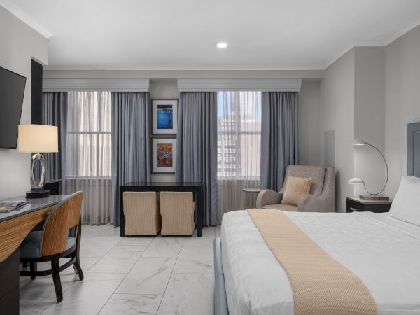 A modern hotel room with a bed, desk, chair, TV, and large windows with curtains, featuring neutral tones and minimalistic decor.
