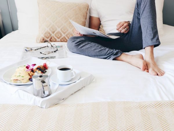A person relaxes on a bed with a breakfast tray holding coffee, a meal, and a magazine, creating a cozy morning scene.