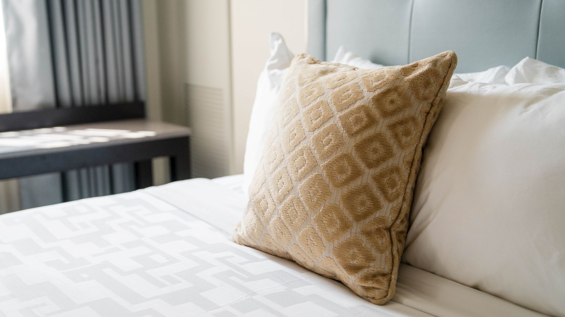 The image shows a neatly made bed with a patterned cushion, white pillows, and a quilt, set in a softly lit room.