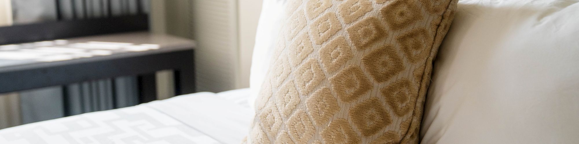 The image shows a neatly made bed with a patterned cushion, white pillows, and a quilt, set in a softly lit room.