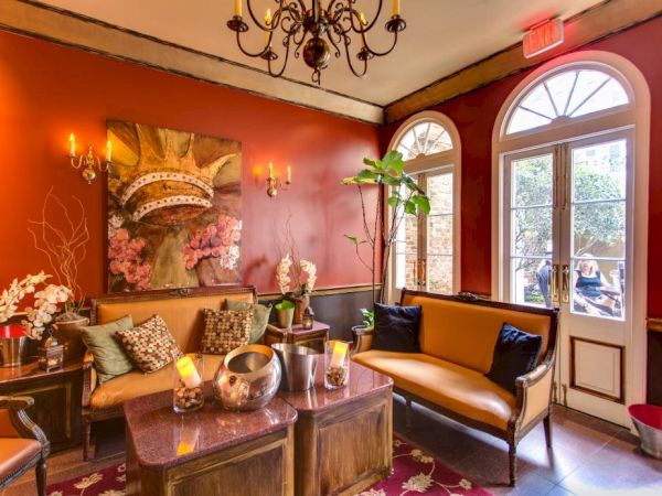 A cozy, elegant lounge with red walls, vintage furniture, chandeliers, and a glass door overlooking a courtyard; plants, candles, and artwork add warmth.