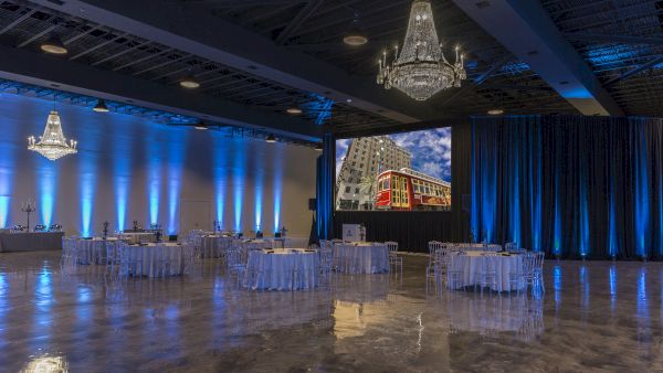 A spacious event hall with chandeliers, elegant tables, blue lighting, and a large screen displaying a city scene with a red streetcar.