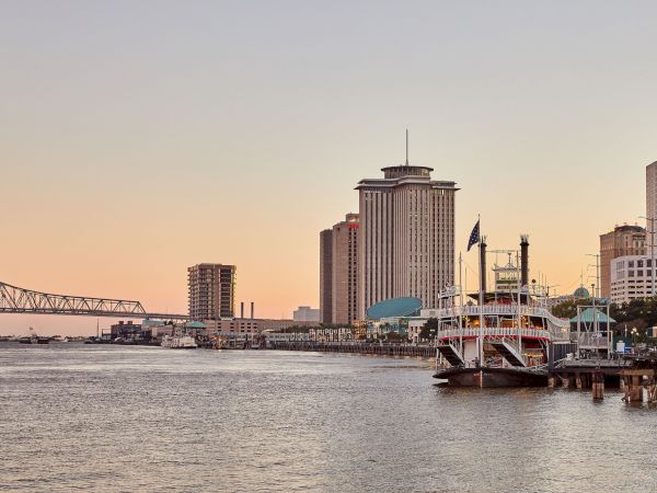 A riverfront skyline at dusk with a tall tower, a large bridge, and a docked cruise ship on the water.