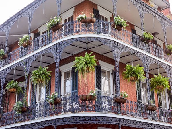 A brick building&rsquo;s balconies with ornate wrought-iron railings, hanging flower pots, and many lush green plants in sunlit doorways.