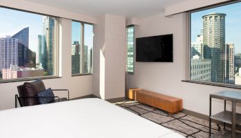 A modern hotel room with large windows showing a city view, a TV, chair, and bed. Urban scenery is visible through the windows.