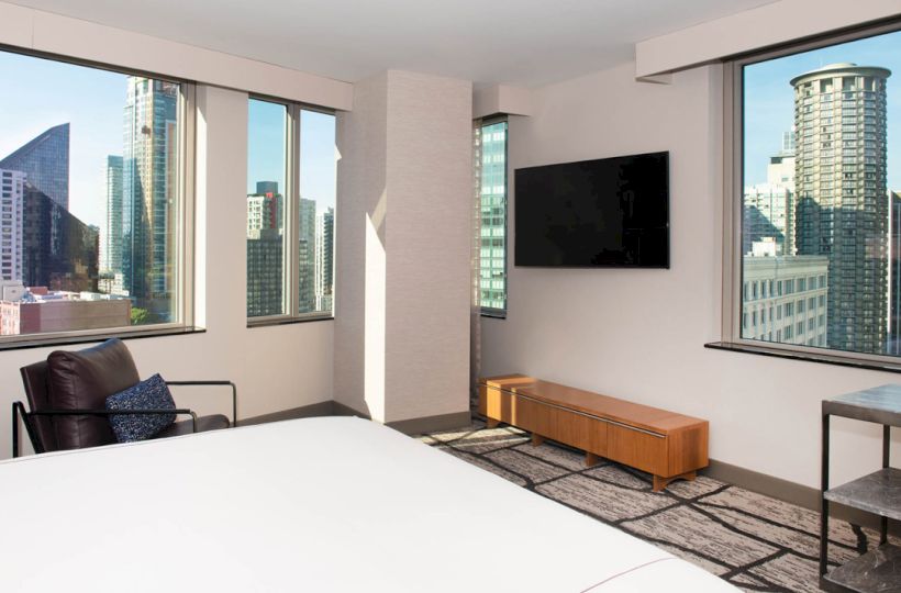 A modern hotel room with large windows showing a city view, a TV, chair, and bed. Urban scenery is visible through the windows.
