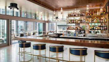 The image shows a modern bar with high stools, a well-stocked liquor shelf, and windows overlooking a cityscape.