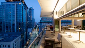 A modern outdoor terrace overlooking a cityscape with tall buildings and a lively street below, featuring seating and open-air space.