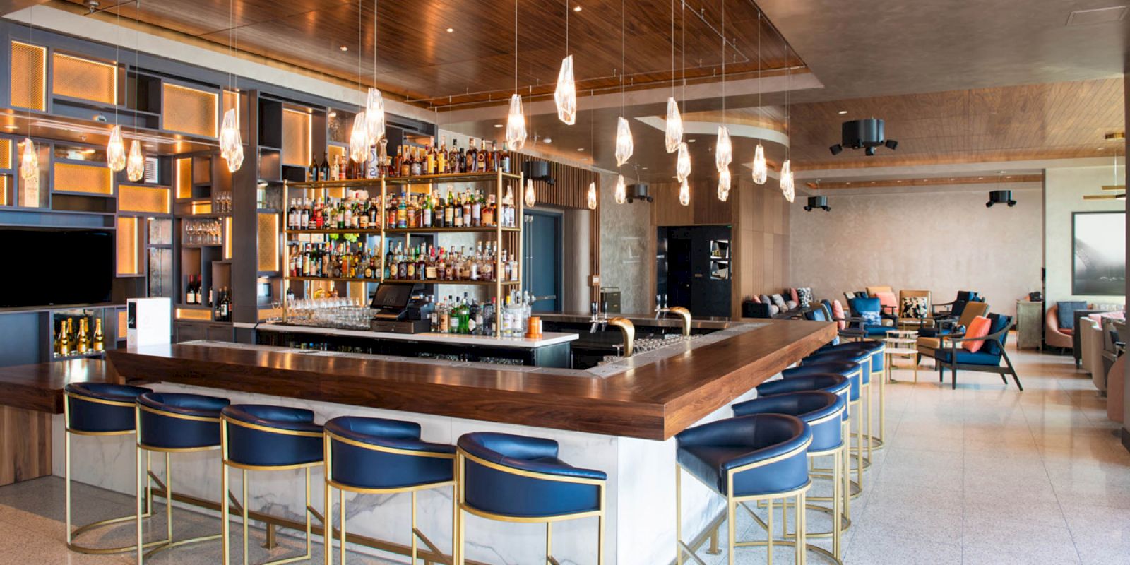 The image shows a modern bar with a central counter, blue chairs, pendant lights, and a variety of bottles displayed.