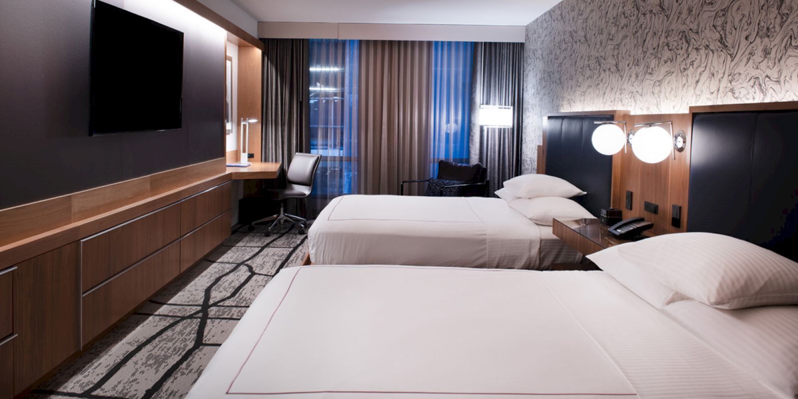 The image shows a modern hotel room with two beds, a wall-mounted TV, a desk with a chair, and stylish decor throughout.