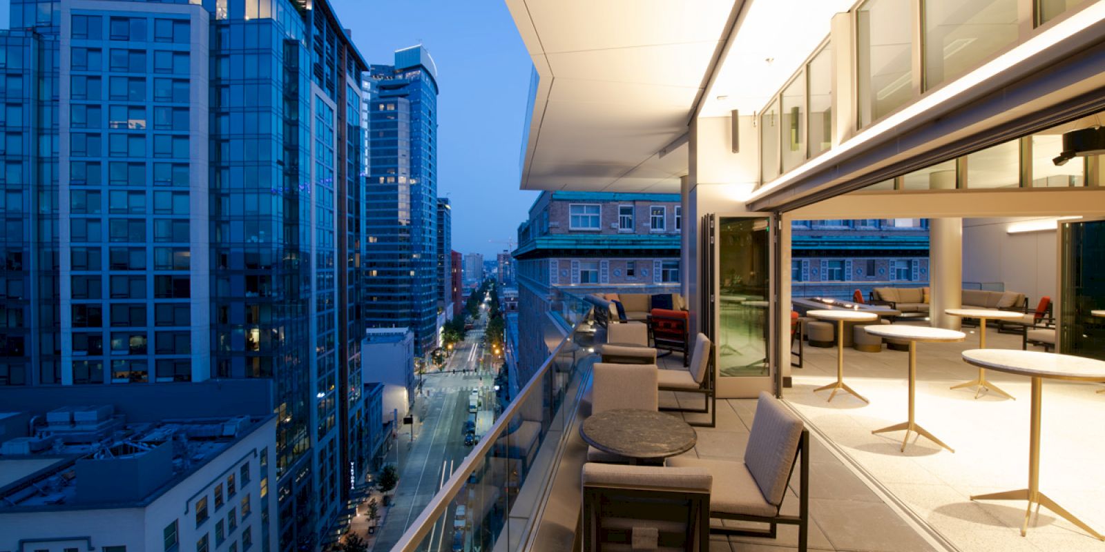 A modern cityscape view from a balcony at dusk, with outdoor seating and tall buildings lining the street below.