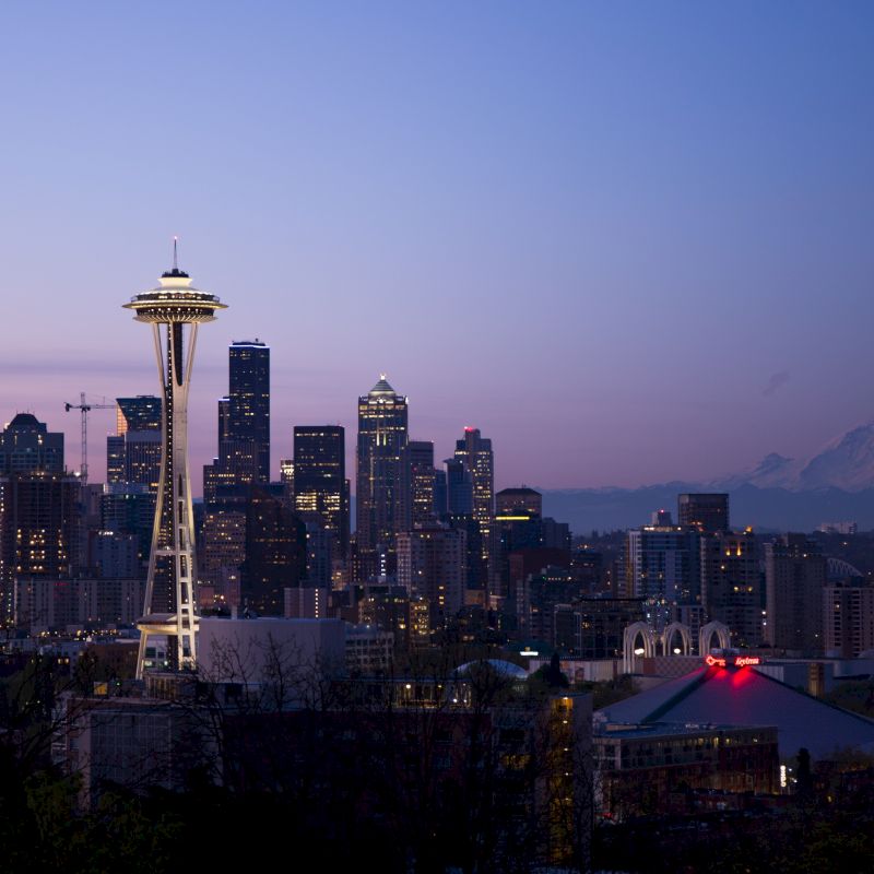 A city skyline at dusk with a prominent tower, skyscrapers, and a mountain in the background under a twilight sky.