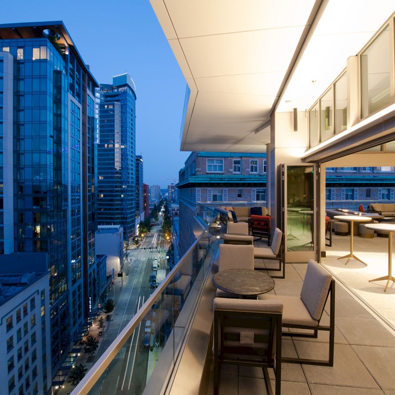 A modern balcony with outdoor seating overlooks a cityscape at dusk, featuring tall buildings and a well-lit street below.
