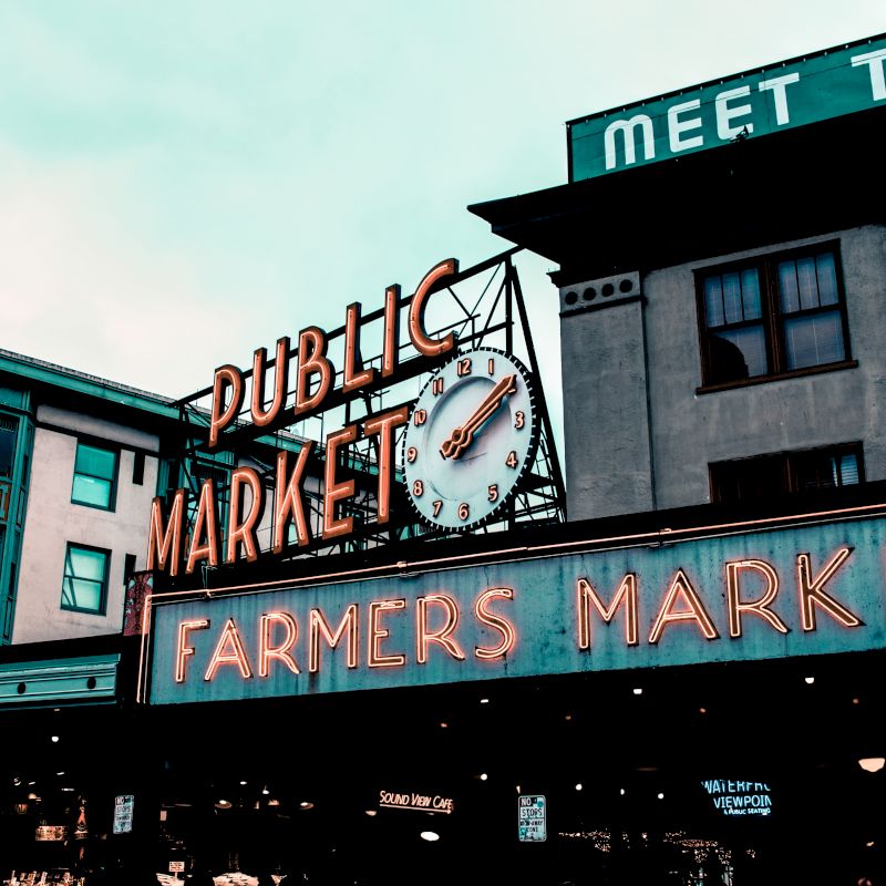 The image shows a neon sign for a public market and farmers market, with buildings in the background and a clock.