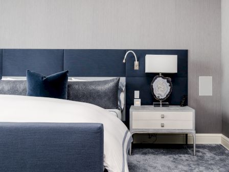 A modern bedroom with a navy bed and white bedding, a nightstand with a lamp, and a decorative marble accent piece beside it.