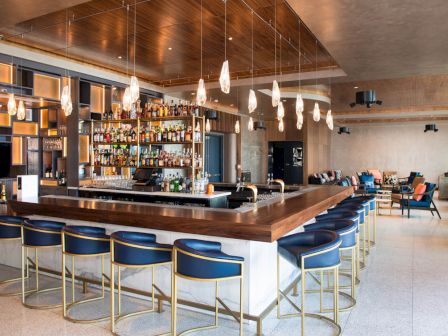 A modern bar with a well-stocked shelf, blue stools, hanging lights, and a cozy seating area in the background.