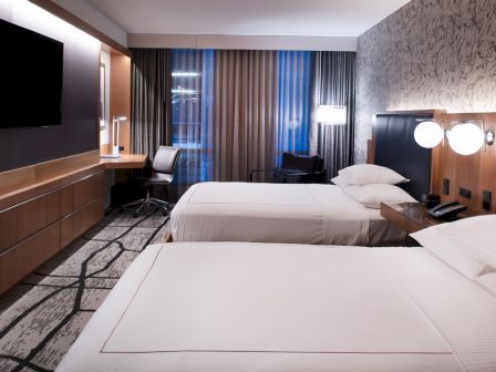 The image shows a modern hotel room with two beds, a desk, a TV, and stylish lighting.