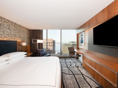 A modern hotel room with a large bed, desk, flat-screen TV, and a view of city buildings through large windows.