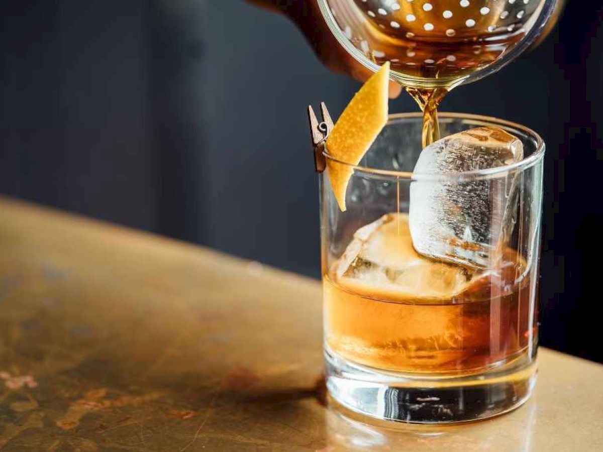 A drink is being poured into a glass with ice and garnished with a lemon twist on a wooden surface.
