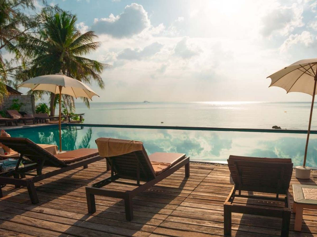 Lounge chairs and umbrellas by a pool overlook a serene ocean view, with palm trees and a partly cloudy sky in the background.