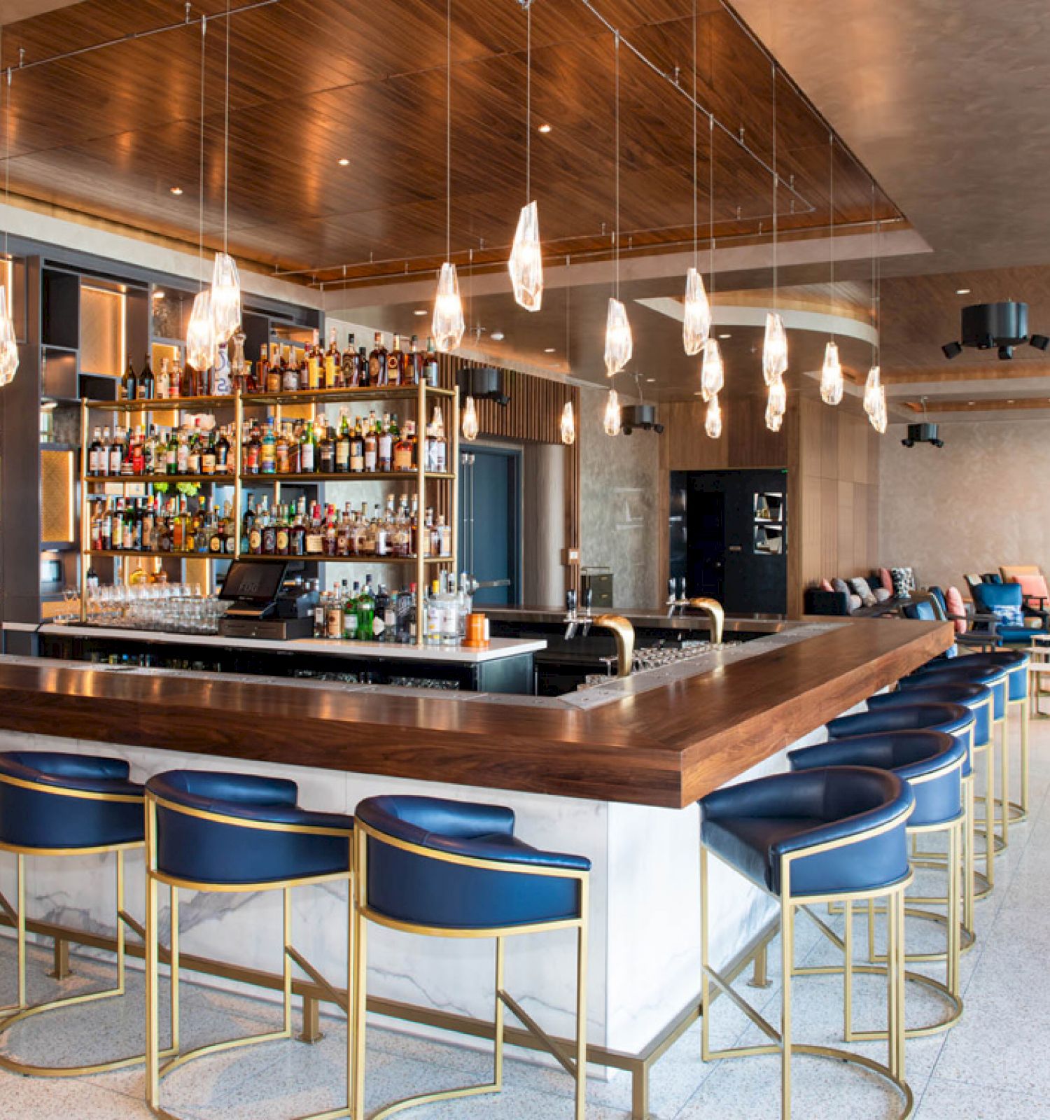 A modern bar with blue stools, a well-stocked shelf, hanging lights, and sleek wooden accents create a stylish and inviting atmosphere.
