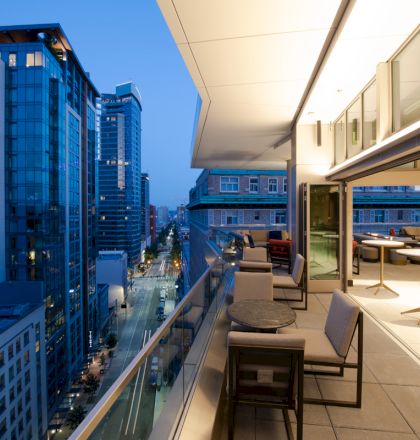 A modern balcony with tables and seating overlooks an urban street at dusk, surrounded by tall buildings under a blue sky.