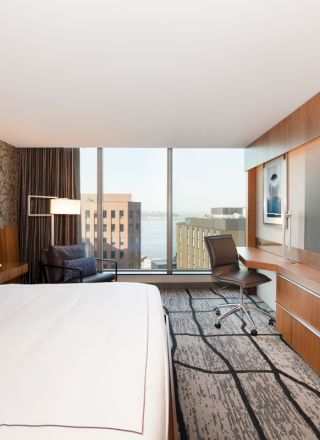 The image shows a modern hotel room with a large bed, a desk, a TV, and a view of buildings and water through large windows.