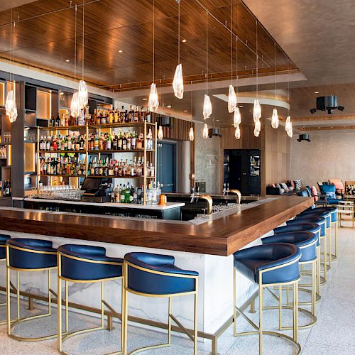 The image shows a modern bar with blue stools, hanging lights, a well-stocked alcohol display, and seating area in the background.