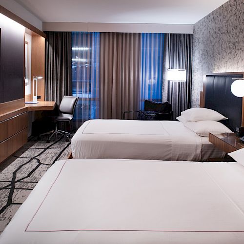 A modern hotel room features two beds, a desk, a large TV, and stylish decor with patterned carpet and wallpaper.