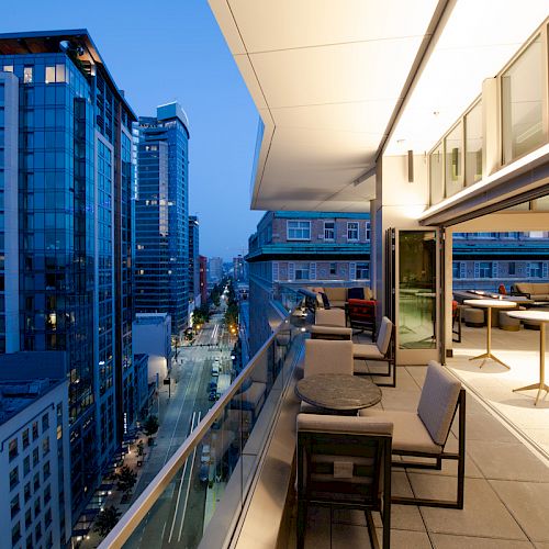 A modern urban balcony overlooking a city street with tall buildings at dusk, featuring seating and tables for relaxation.