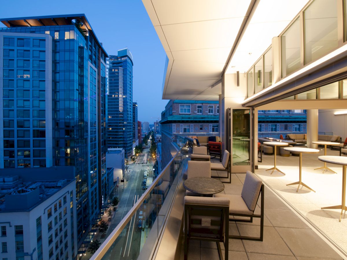 A modern outdoor balcony with tables and seating overlooks a cityscape of tall buildings during the evening.