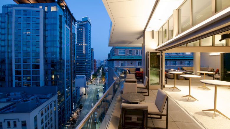 A modern outdoor balcony with tables and seating overlooks a cityscape of tall buildings during the evening.