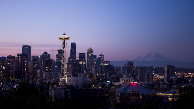 The image shows a skyline view of Seattle at dusk, featuring the Space Needle and Mount Rainier in the background.