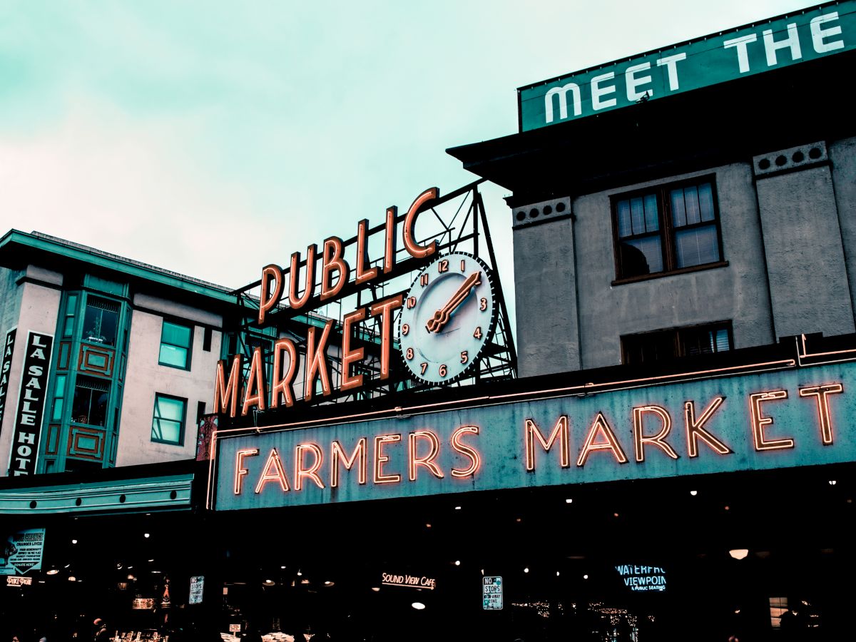 A market sign reads "Public Market" and "Farmers Market" with a clock, on buildings beneath a cloudy sky.