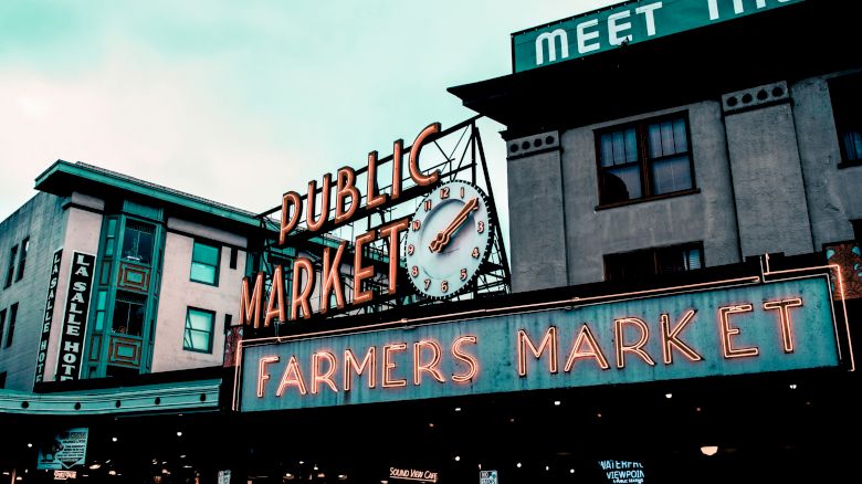 A market sign reads "Public Market" and "Farmers Market" with a clock, on buildings beneath a cloudy sky.