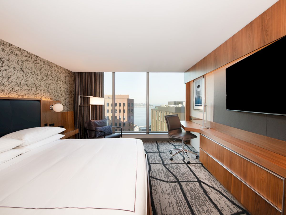 A modern hotel room with a large bed, desk, chair, TV, and window view of buildings and water is shown.