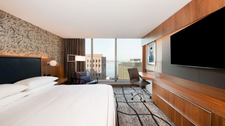 A modern hotel room with a large bed, desk, chair, TV, and window view of buildings and water is shown.