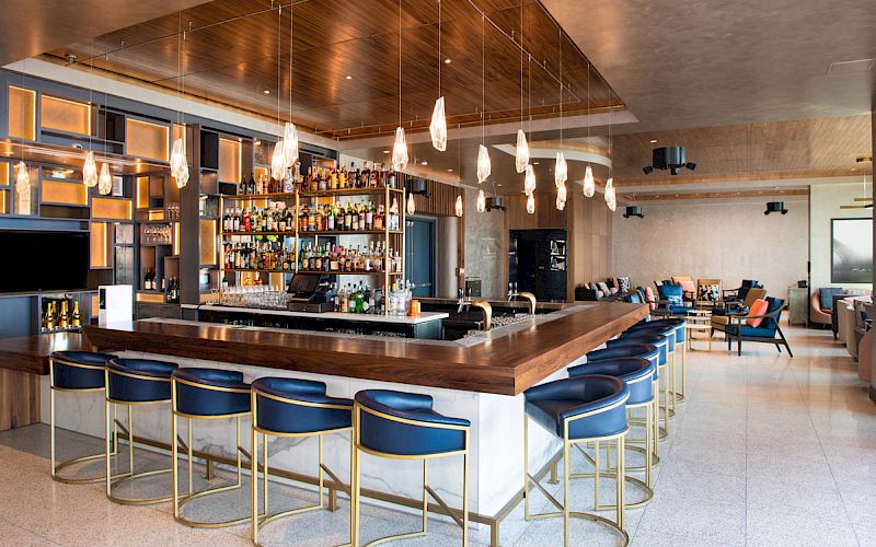 A modern bar with blue stools, wooden accents, pendant lights, and a stocked backlit bar, leading to a cozy seating area further back.
