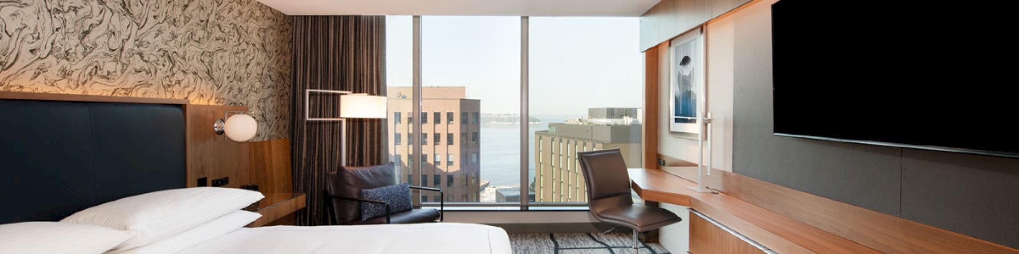 A modern hotel room with a large bed, a wall-mounted TV, a chair, and a desk, overlooking city buildings and water through the window.